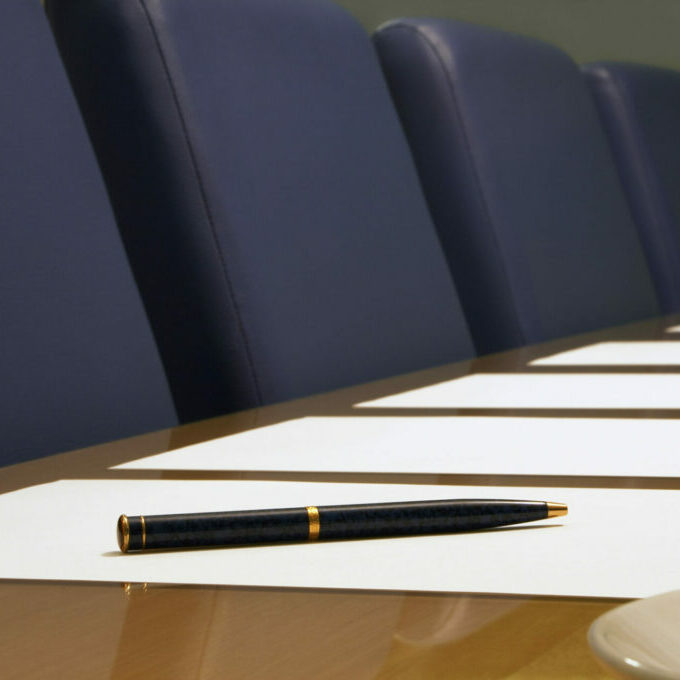 A close-up of a conference room showing a row of chairs, a table, part of a coffee cup, documents (blank) and a pen resting on a piece of paper. Sharp focus on the pen.